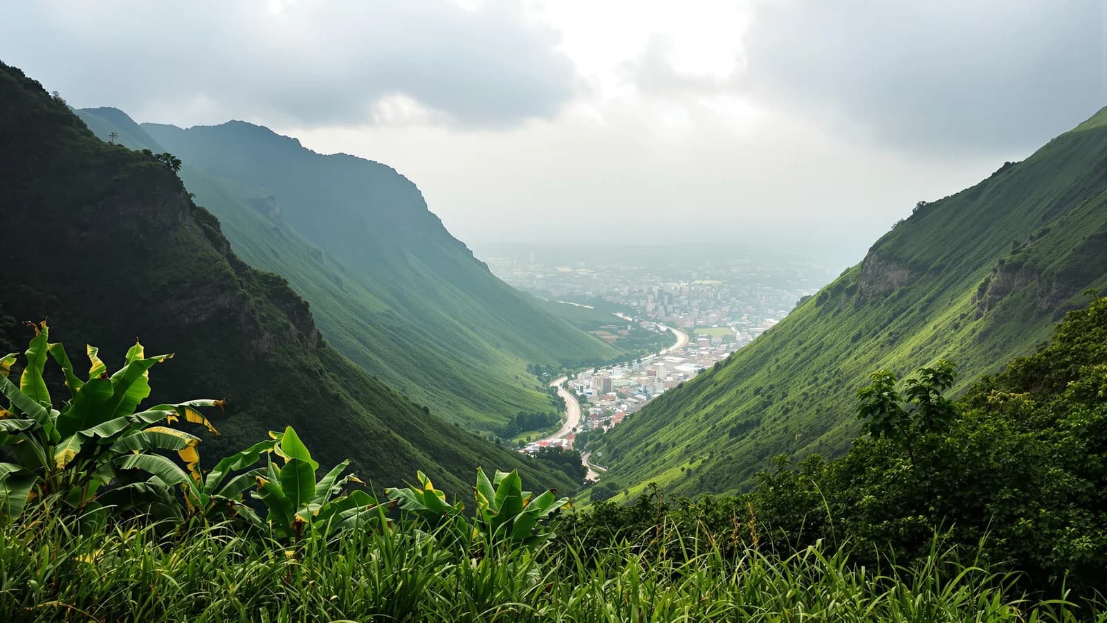 Steile grüne Hänge rahmen ein langes Tal mit einer Stadt am Talgrund, Wolkenschatten auf den Hängen.