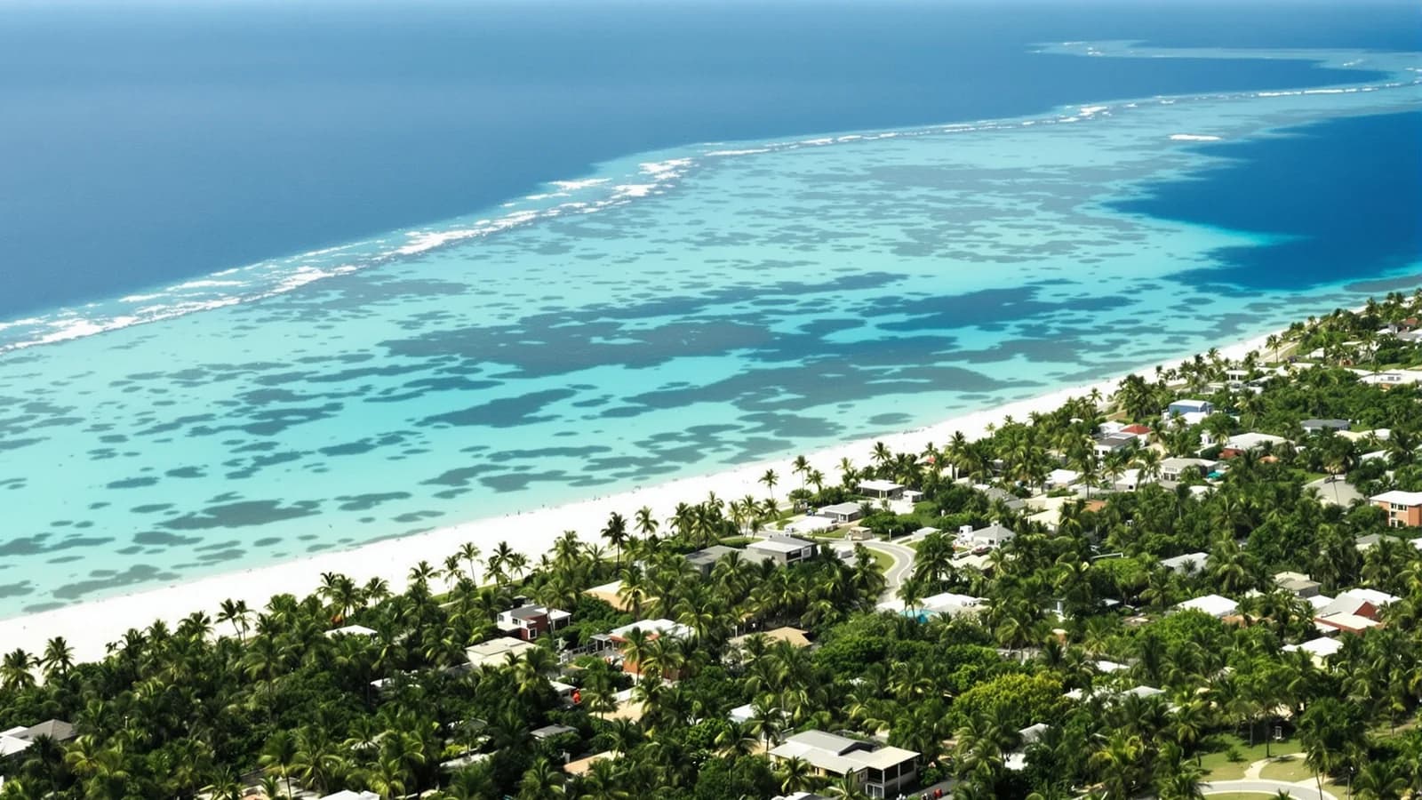 Karibikküste aus der Höhe, weißer Strand, flache Dächer zwischen Palmen, türkis gebändertes Meer.